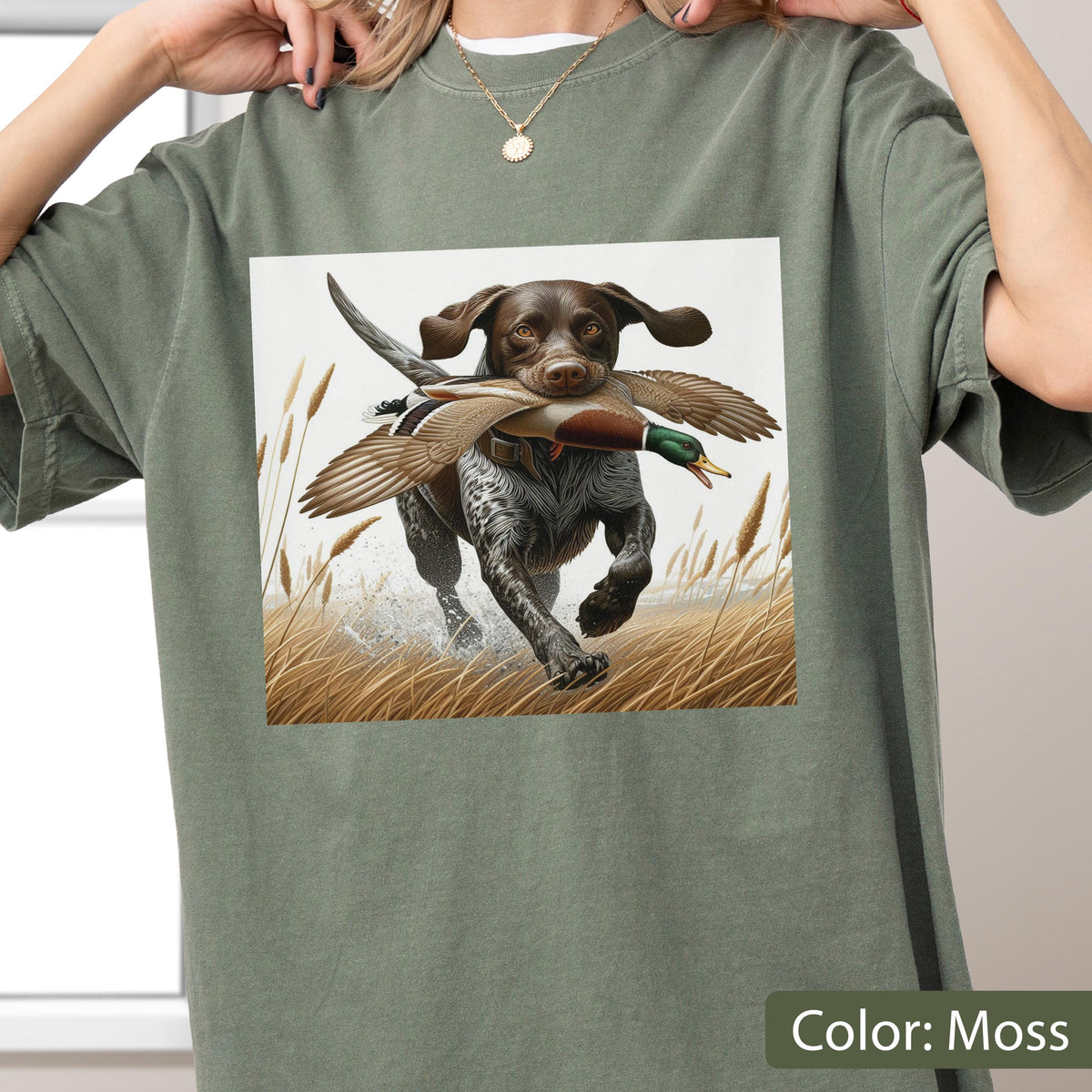 May include: A moss green t-shirt featuring a graphic print of a brown and white spotted hunting dog running through tall golden grass while carrying a mallard duck in its mouth. The dog has floppy ears and is depicted in a field setting.