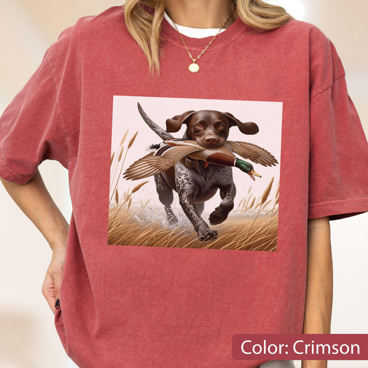 May include: A crimson short-sleeve t-shirt featuring a graphic print of a brown and white hunting dog running through tall grass while carrying a mallard duck in its mouth. The dog has floppy ears and a spotted coat, set against a light background.