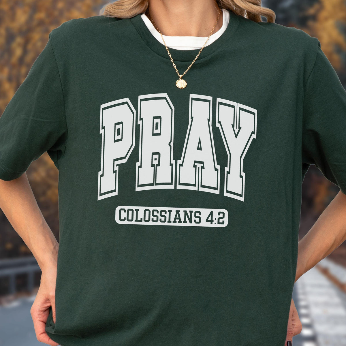 May include: A dark forest green short-sleeve t-shirt featuring the word PRAY in large, bold, light gray block letters across the chest. Below the word, the text COLOSSIANS 4:2 is printed in a smaller, matching light gray font on a white background.