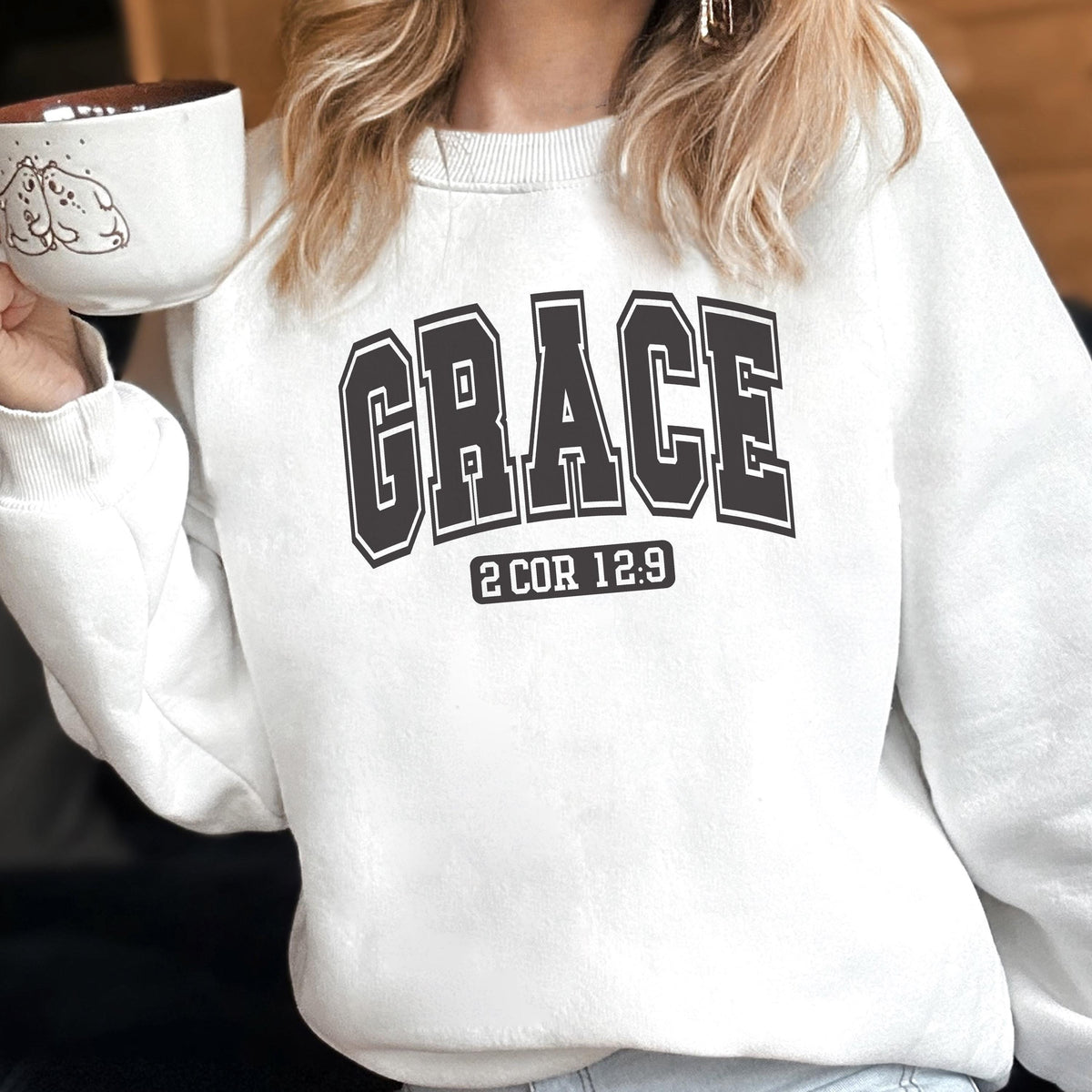 May include: A white crewneck sweatshirt featuring the word GRACE in a large, dark, collegiate-style font across the chest. Below the word, the text 2 COR 12:9 is printed in a smaller, matching dark font. The sweatshirt has long sleeves and ribbed cuffs.