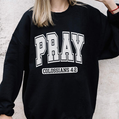 May include: A black long-sleeve sweatshirt featuring the word PRAY in large, white, collegiate-style block lettering across the chest. Below the word PRAY, the text COLOSSIANS 4:2 is printed in a smaller, white, rectangular block font.