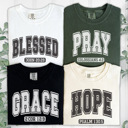 May include: Four t-shirts in white, dark green, black, and cream. Each shirt features collegiate-style lettering and a Bible verse: BLESSED JOHN 20:29, PRAY COLOSSIANS 4:2, GRACE 2 COR 12:9, and HOPE PSALM 130:5. All shirts have a neck label.