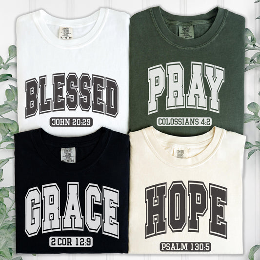 May include: Four t-shirts in white, dark green, black, and cream. Each shirt features collegiate-style lettering and a Bible verse: BLESSED JOHN 20:29, PRAY COLOSSIANS 4:2, GRACE 2 COR 12:9, and HOPE PSALM 130:5. All shirts have a neck label.