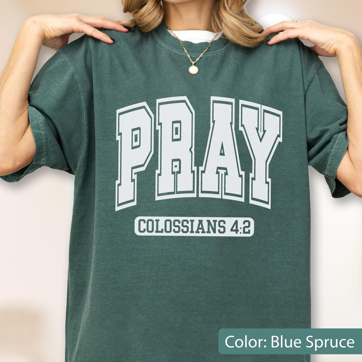May include: A blue spruce short-sleeve t-shirt featuring the word PRAY in large white collegiate-style block letters across the chest. Below the word PRAY, the text COLOSSIANS 4:2 is printed in a smaller white font within a rectangular border.