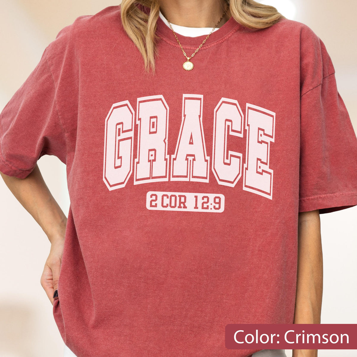 May include: A crimson short-sleeve t-shirt featuring the word GRACE in large, light pink collegiate-style block letters. Below the word, the text 2 COR 12:9 is printed in a smaller, matching light pink font. The shirt has a crew neckline.