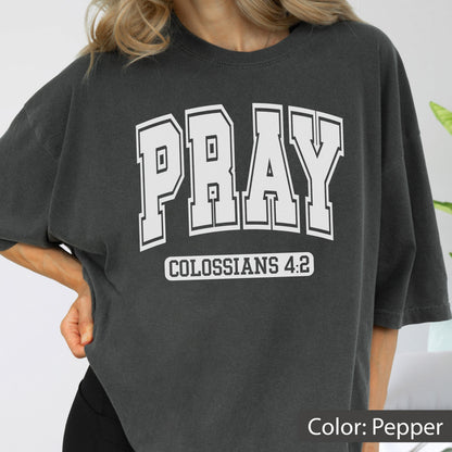 May include: A pepper-colored short-sleeve t-shirt featuring the word PRAY in large white collegiate-style block letters. Below the word PRAY, the text COLOSSIANS 4:2 is printed in a smaller white rectangular box with a clean sans-serif font.