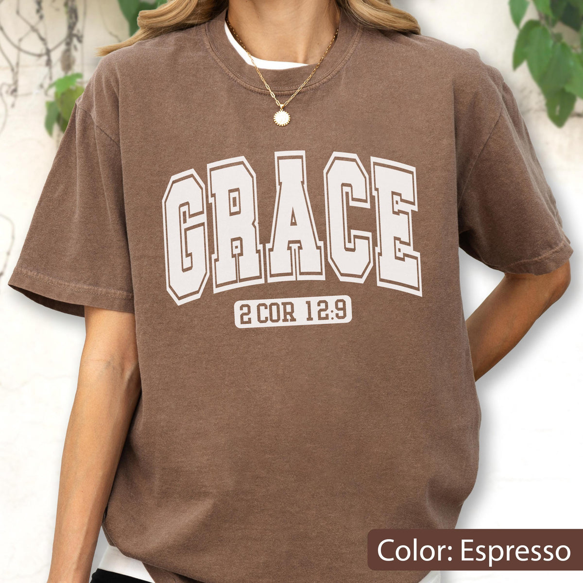 May include: An espresso brown short-sleeve t-shirt featuring the word GRACE in large, white, collegiate-style block lettering across the chest, with the text 2 COR 12:9 printed in a smaller white font directly underneath the main word.