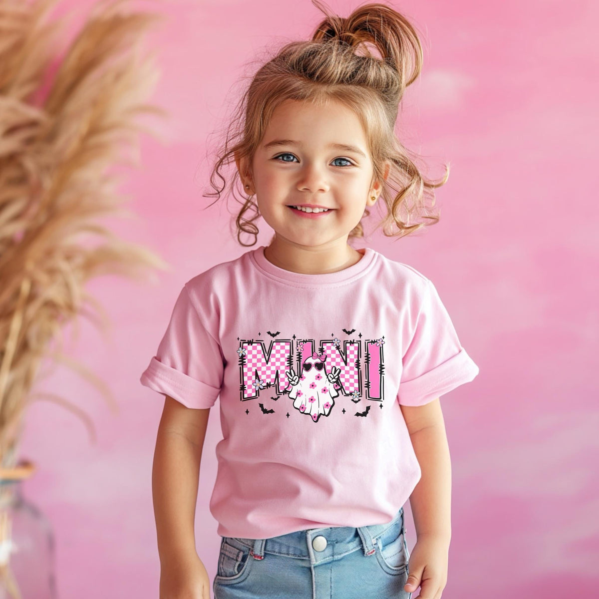 May include: A light pink short-sleeved t-shirt featuring a graphic print with the word MINI in a pink and white checkered pattern. The design includes a white ghost character wearing pink sunglasses and floral accents, surrounded by small black bats.