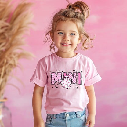 May include: A light pink short-sleeved t-shirt featuring a graphic print with the word MINI in a pink and white checkered pattern. The design includes a white ghost character wearing pink sunglasses and floral accents, surrounded by small black bats.
