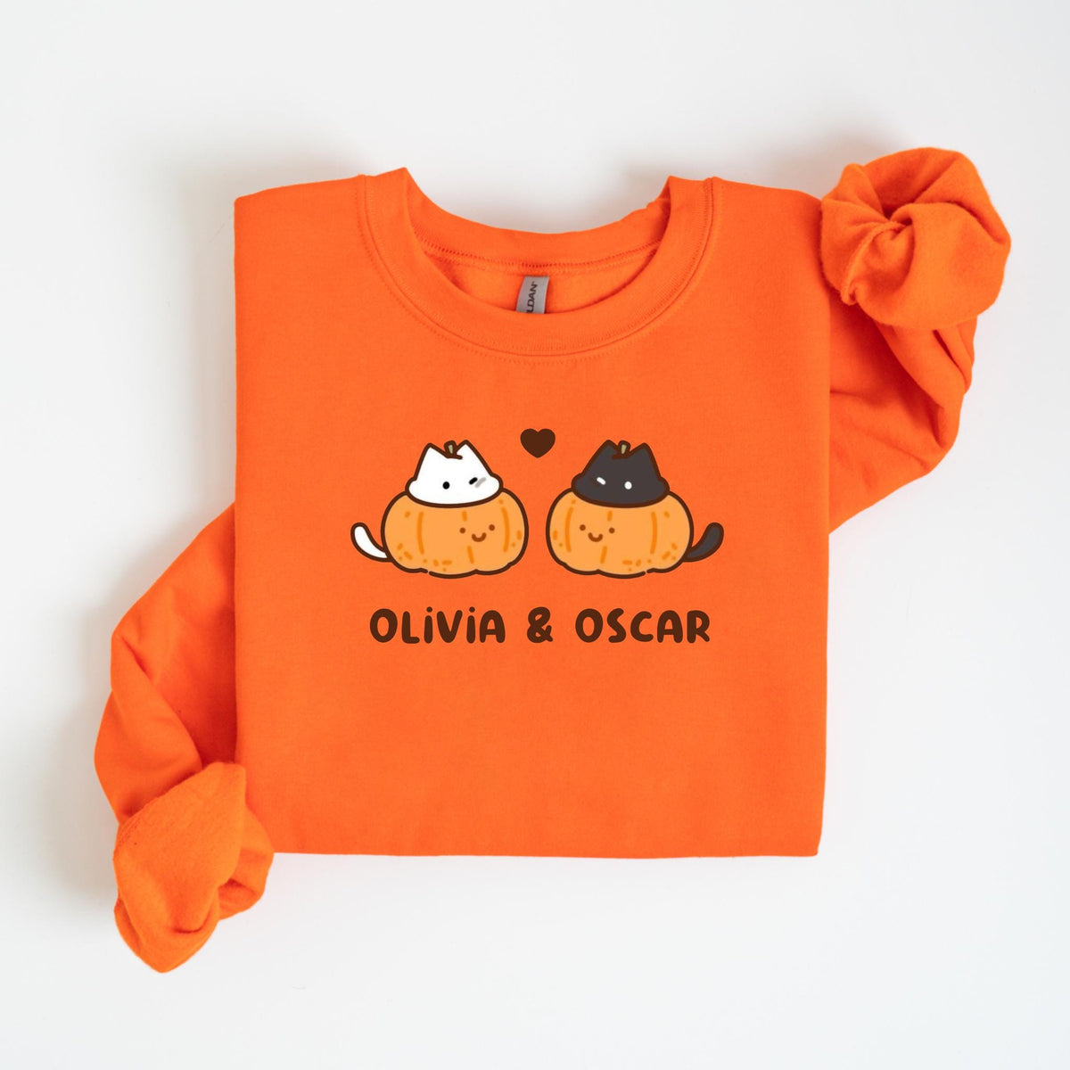 May include: Orange sweatshirt with a crew neck. The front features a graphic of two cats inside pumpkins, with a heart above them. The text "OLIVIA & OSCAR" is printed below the graphic. The sleeves are long.