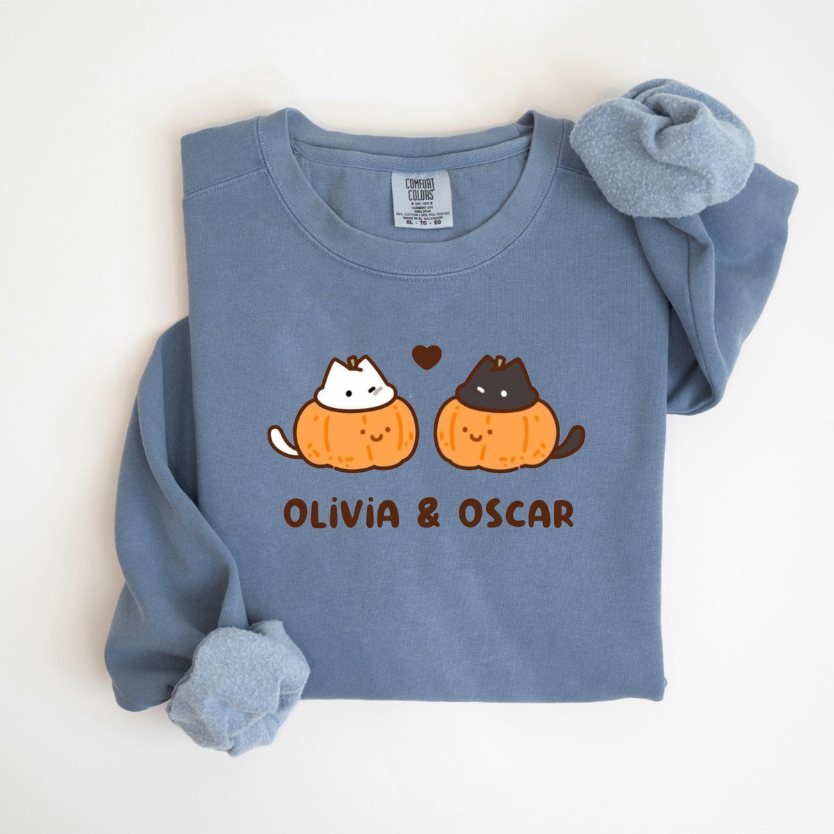May include: A blue long-sleeve sweatshirt featuring a cartoon design of two cats inside orange pumpkins with the text "Olivia & Oscar" below. The cats are a white cat and a black cat. A small brown heart is above the cats.