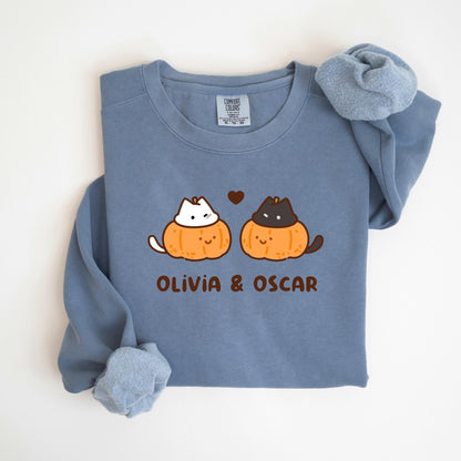 May include: A blue long-sleeve sweatshirt featuring a cartoon design of two cats inside orange pumpkins with the text "Olivia & Oscar" below. The cats are a white cat and a black cat. A small brown heart is above the cats.