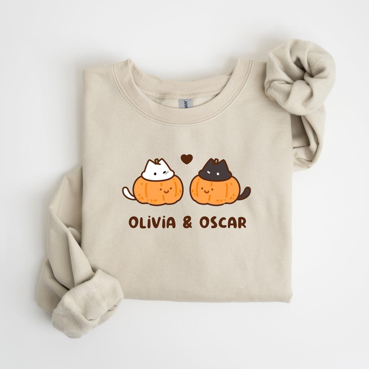 May include: Beige sweatshirt featuring a graphic of two cartoon cats inside orange pumpkins, with a small heart above them. The text "OLIVIA & OSCAR" is printed below the pumpkins. The sweatshirt has long sleeves and a crew neck.