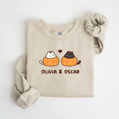 May include: Beige sweatshirt featuring a graphic of two cartoon cats inside orange pumpkins, with a small heart above them. The text "OLIVIA & OSCAR" is printed below the pumpkins. The sweatshirt has long sleeves and a crew neck.