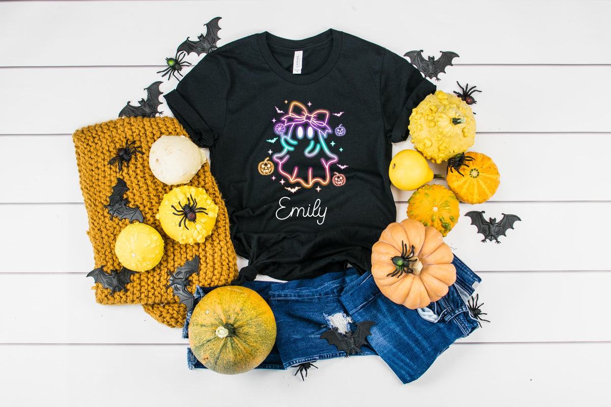 May include: Black t-shirt with a neon ghost design, the name "Emily" in white script, and Halloween decorations. The shirt is paired with denim shorts, pumpkins, and a knitted scarf.