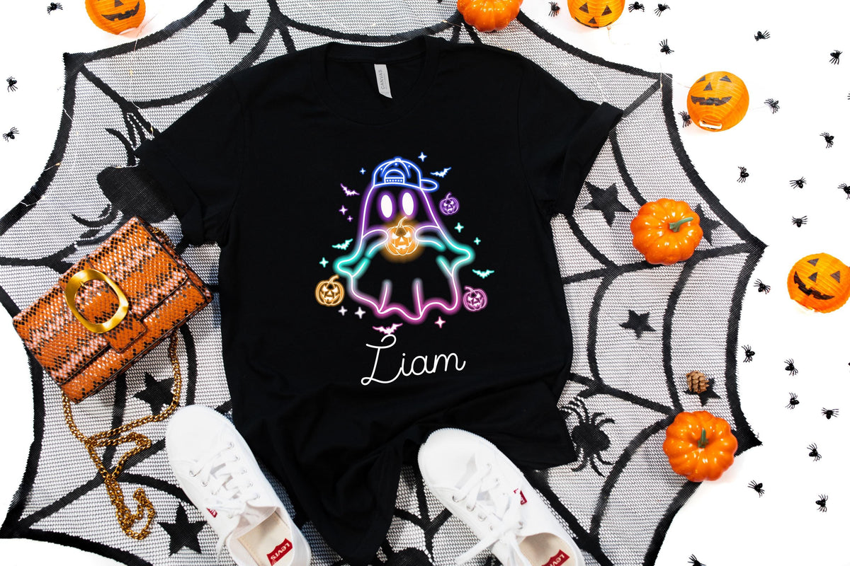 May include: Black t-shirt featuring a neon ghost design with a baseball cap and holding a pumpkin. The name "Liam" is written in cursive. The shirt is surrounded by Halloween decorations, including pumpkins and a spiderweb.