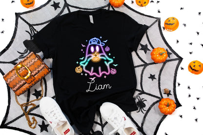 May include: Black t-shirt featuring a neon ghost design with a baseball cap and holding a pumpkin. The name "Liam" is written in cursive. The shirt is surrounded by Halloween decorations, including pumpkins and a spiderweb.