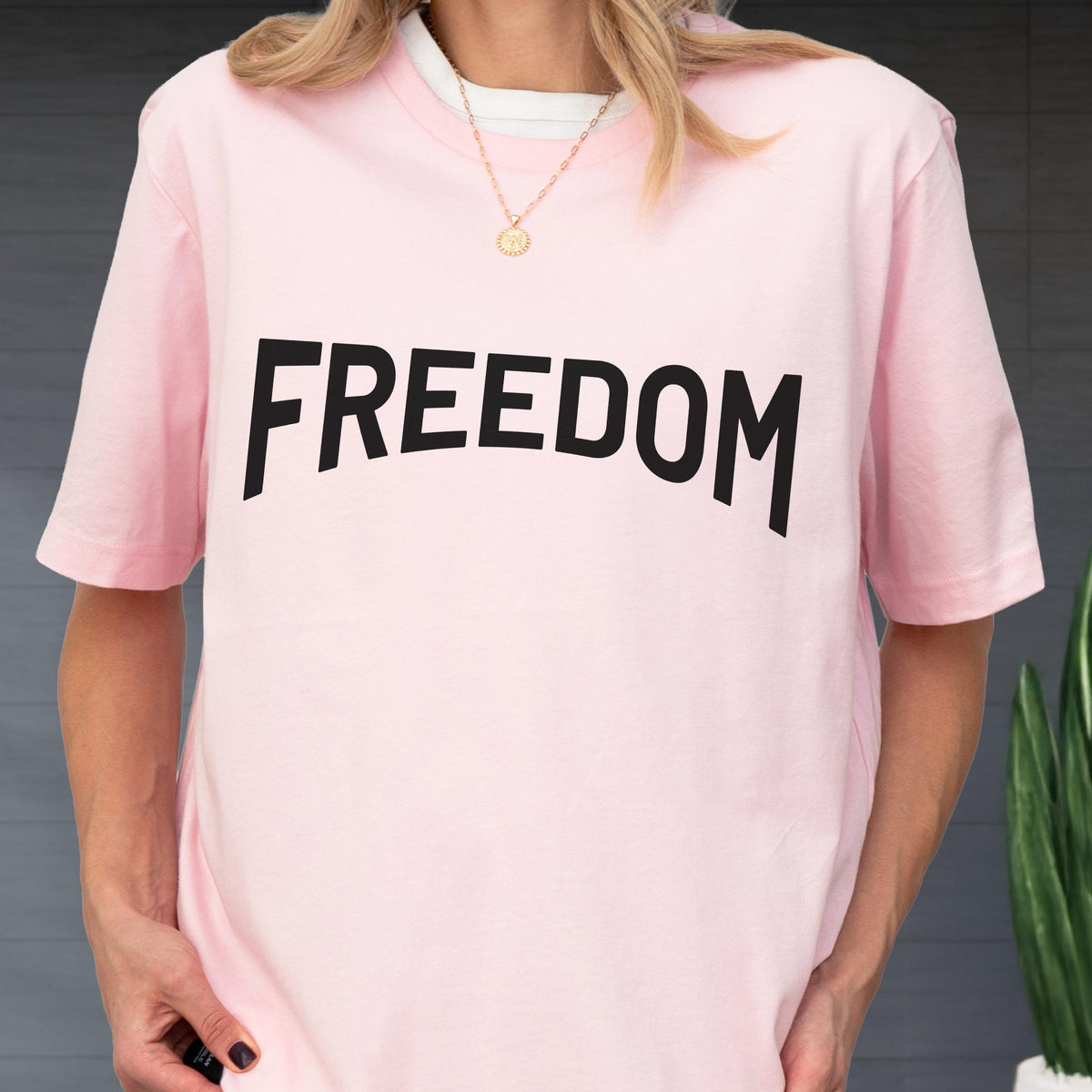 May include: A light pink t-shirt with the word "FREEDOM" printed in large, black, bold letters across the front. The shirt has short sleeves and a crew neck. A gold necklace is visible.