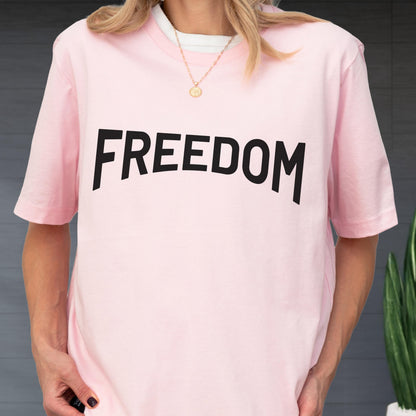 May include: A light pink t-shirt with the word "FREEDOM" printed in large, black, bold letters across the front. The shirt has short sleeves and a crew neck. A gold necklace is visible.