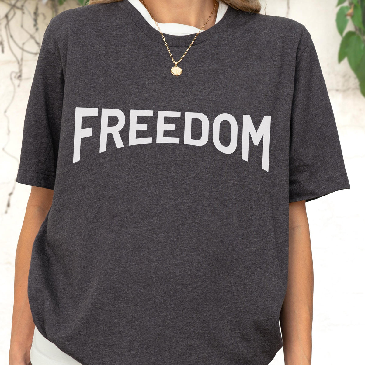 May include: Dark gray t-shirt with the word "FREEDOM" printed in large, white capital letters across the chest. The shirt has a white collar and short sleeves. A gold necklace with a pendant is visible.