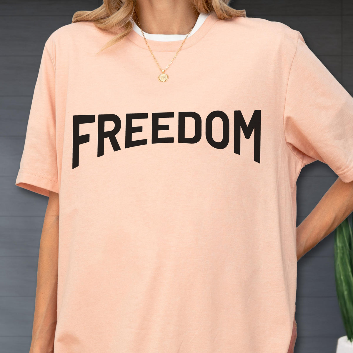 May include: A light peach-colored t-shirt with the word "FREEDOM" printed in bold black letters. The shirt has a white crew neck collar and short sleeves. A gold necklace with a pendant is visible.