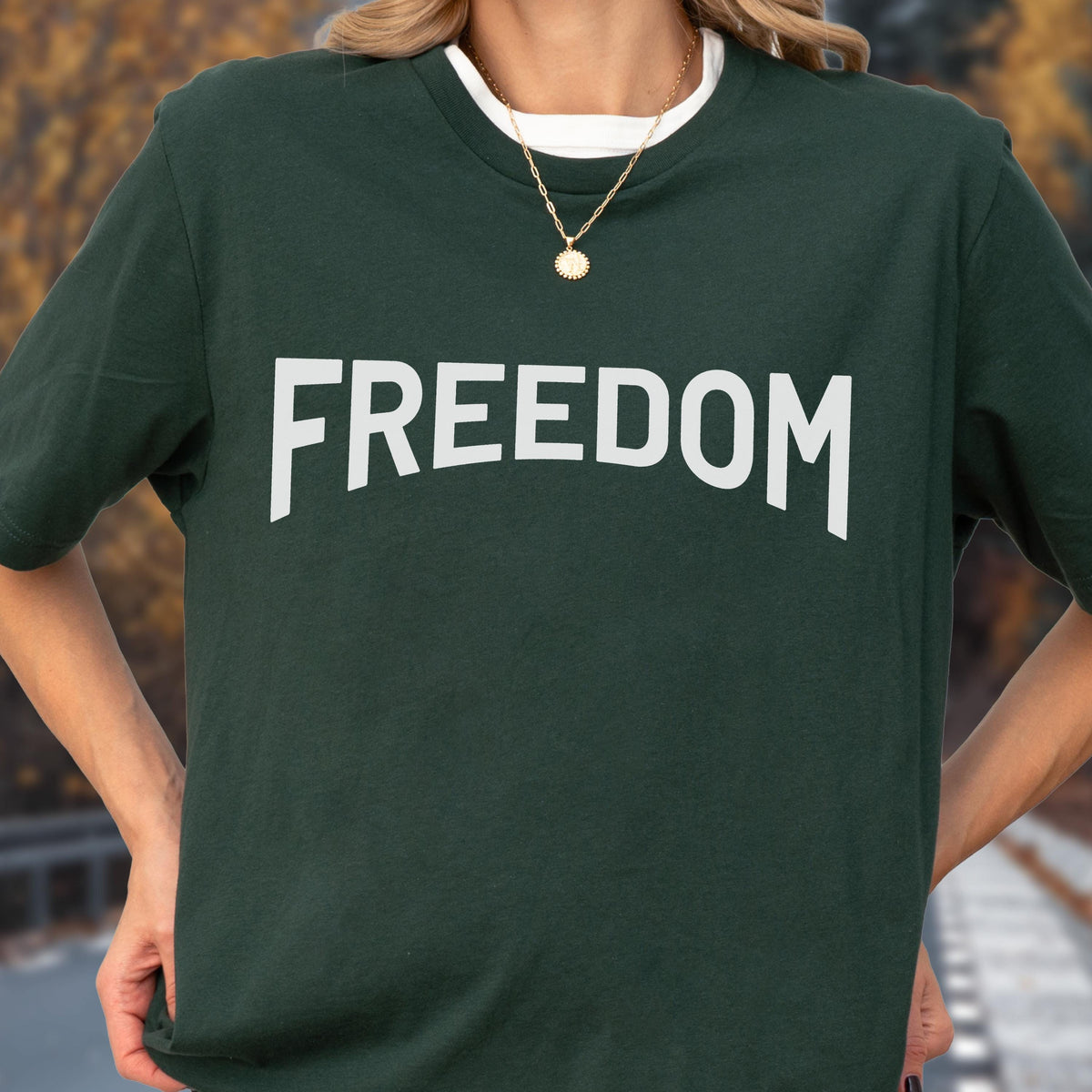 May include: Dark green t-shirt with the word "FREEDOM" in large, white, block letters. The shirt has a white crew neck collar and is worn with a gold necklace. The background is blurred, suggesting an outdoor setting.