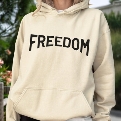 May include: A cream-colored hoodie with the word "FREEDOM" printed in bold, black letters across the chest. The hoodie has a front pocket and a hood. The background is blurred, suggesting an outdoor setting.