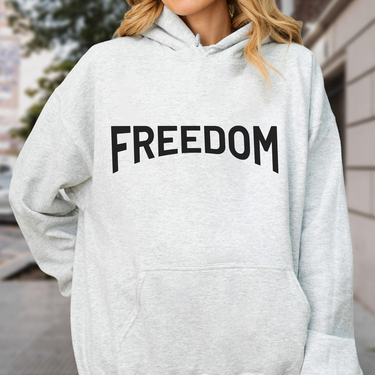 May include: A light gray hoodie with the word "FREEDOM" printed in bold black letters across the chest. The hoodie has a front pocket and a hood. The background is blurred, suggesting an outdoor setting.