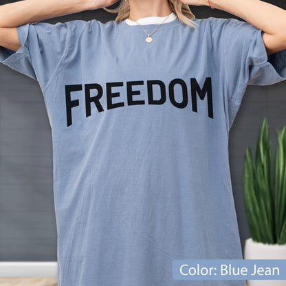 May include: Blue jean colored t-shirt with the word "FREEDOM" printed in large, black letters across the front. The shirt has short sleeves and a crew neck. The shirt is a solid color.