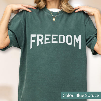 May include: A blue spruce-colored t-shirt with the word "FREEDOM" printed in large, white, block letters. The shirt has short sleeves and a crew neck. The color is described as "Blue Spruce".