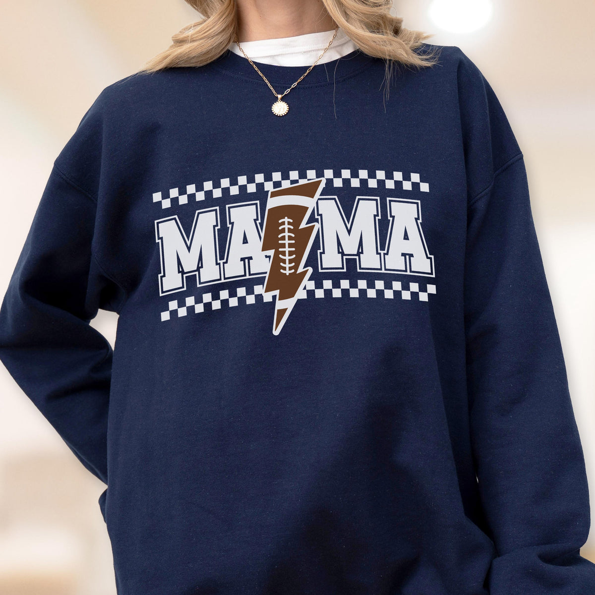 May include: Navy blue sweatshirt with the word "MAMA" in white block letters, a brown football-shaped lightning bolt, and a white and black checkered border. The sweatshirt is a casual style.
