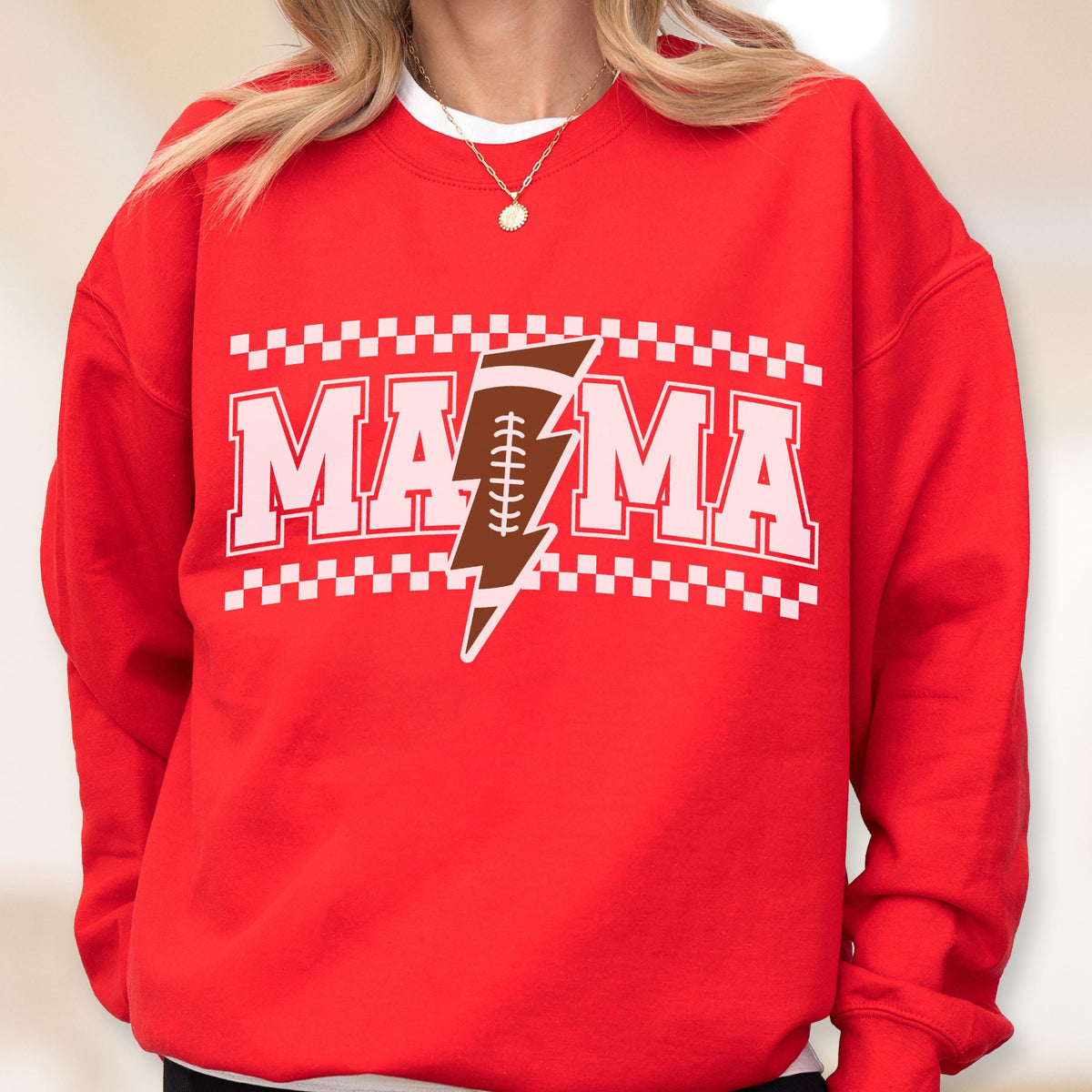 May include: Red sweatshirt with the word "MAMA" in white block letters, with a brown football and lightning bolt graphic. The design is framed by a white and black checkered pattern. A casual, sporty top.