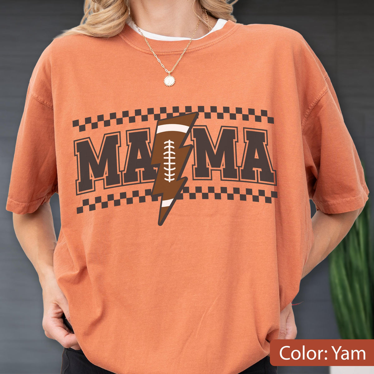 May include: A yam-colored short-sleeve t-shirt featuring a graphic print with the text MAMA in dark brown block letters. A brown lightning bolt shaped like an American football with white laces sits between the letters. A checkered border frames the text.