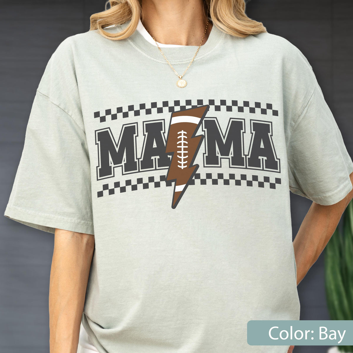 May include: A light sage green short-sleeve t-shirt featuring the text MAMA in black block letters. A brown football-shaped lightning bolt replaces the letter A. The design is framed by black and white checkered borders above and below the text.