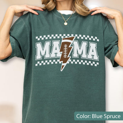 May include: A blue spruce short-sleeve t-shirt featuring the white text MAMA with a brown football shaped like a lightning bolt replacing the letter A. The design is framed by white checkered borders above and below the lettering.
