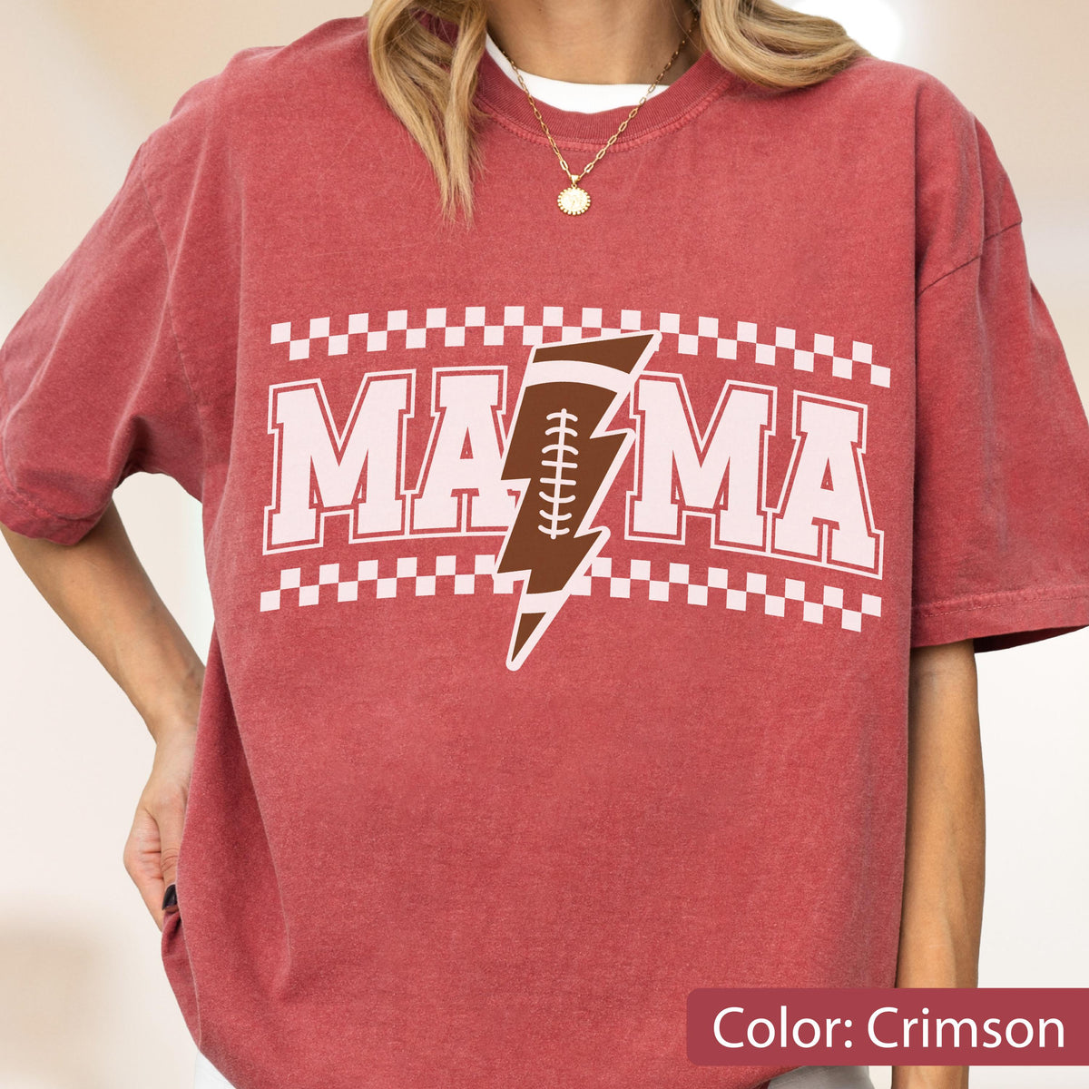 May include: A crimson short-sleeve t-shirt featuring the text MAMA in white block letters. A brown football-shaped lightning bolt graphic is positioned between the two M's. The design is framed by white checkered borders at the top and bottom.