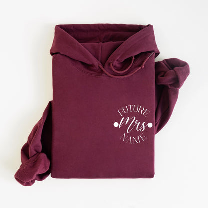 May include: A maroon hoodie with a drawstring hood and a front pocket. The hoodie features the text "FUTURE Mrs NAME" in a white, circular design. The sleeves are folded, and the background is white.