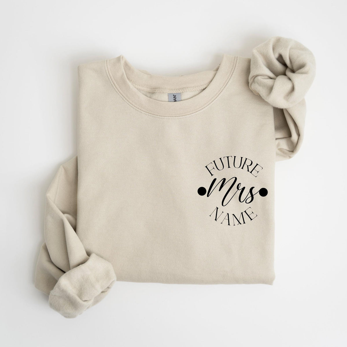 May include: A beige sweatshirt with the text "FUTURE Mrs NAME" in black script. A matching beige scrunchie is placed on the upper right. The sweatshirt is folded, with the sleeves visible.