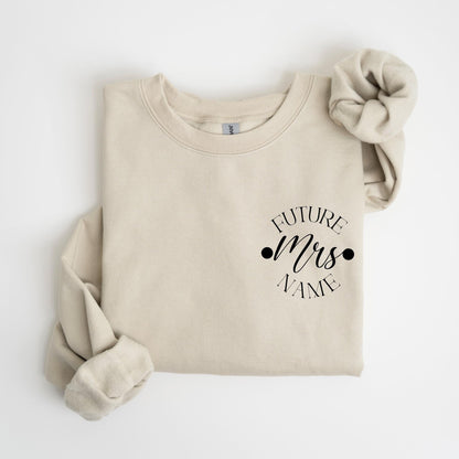 May include: A beige sweatshirt with the text "FUTURE Mrs NAME" in black script. A matching beige scrunchie is placed on the upper right. The sweatshirt is folded, with the sleeves visible.