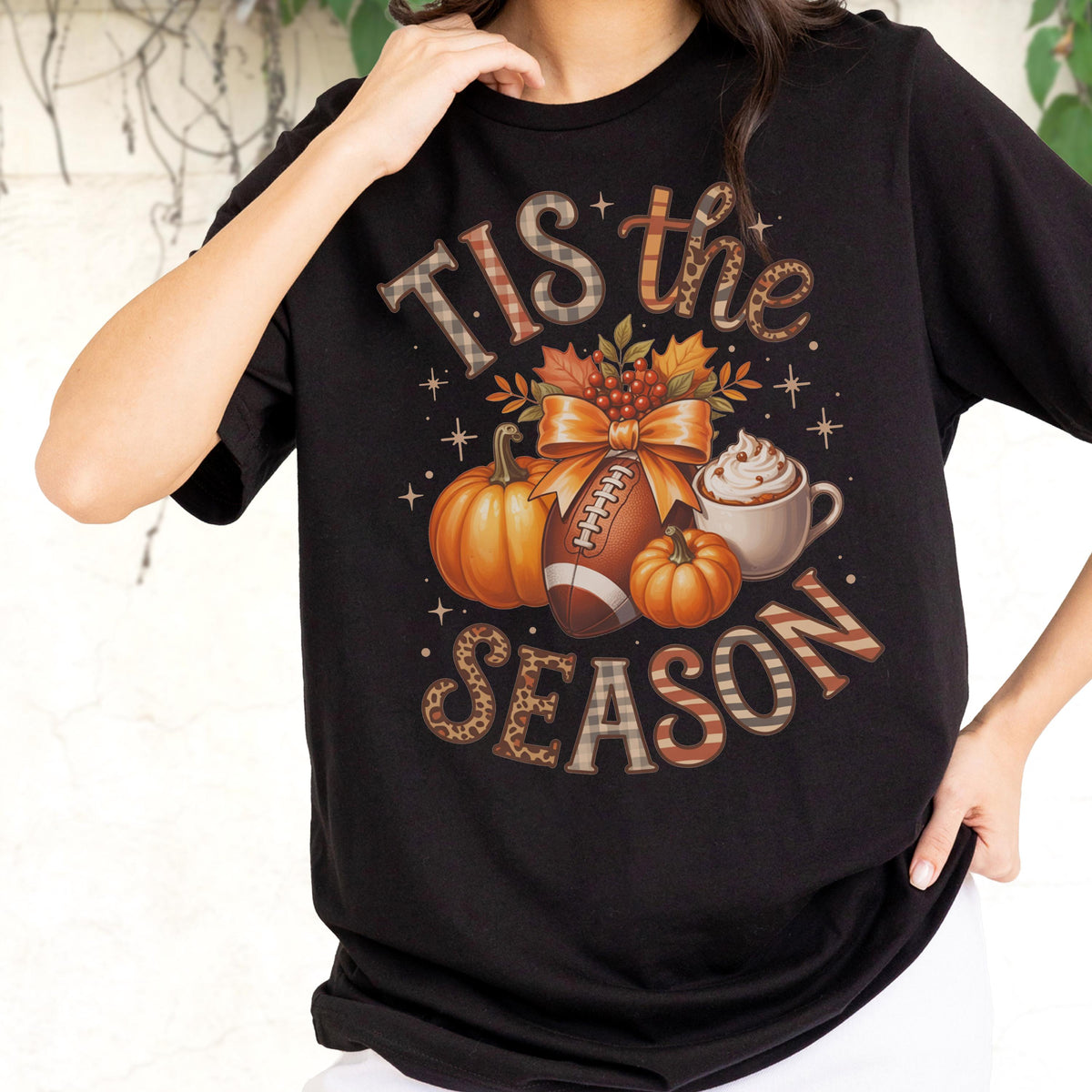 May include: Black t-shirt with a fall-themed graphic. The design includes pumpkins, a football, a bow, and a cup of whipped cream. The text reads "Tis the Season" in various decorative fonts and patterns.