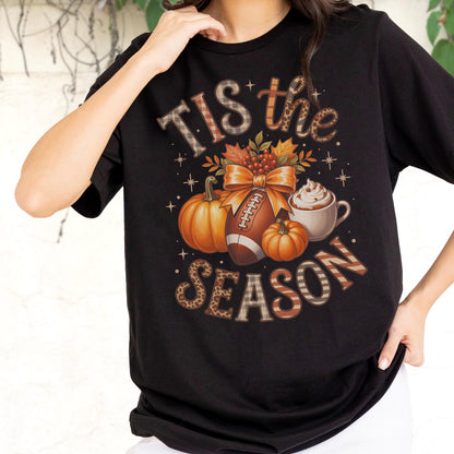 May include: Black t-shirt with a fall-themed graphic. The design includes pumpkins, a football, a bow, and a cup of whipped cream. The text reads "Tis the Season" in various decorative fonts and patterns.