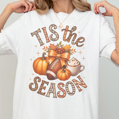 May include: White t-shirt with a fall-themed graphic. The design includes pumpkins, a football, a cup of whipped cream, and the text "TIS the SEASON" in decorative lettering. The overall color scheme is warm, featuring orange, brown, and cream tones.