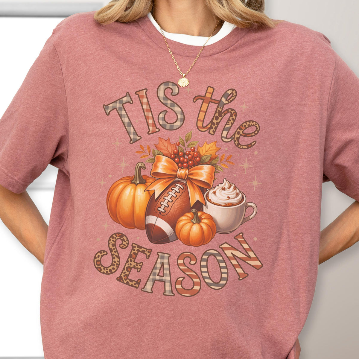 May include: A mauve-colored t-shirt with a fall-themed graphic. The design includes pumpkins, a football, a cup of whipped cream, and the text "TIS the SEASON" in decorative fonts. The shirt has a crew neck and short sleeves.
