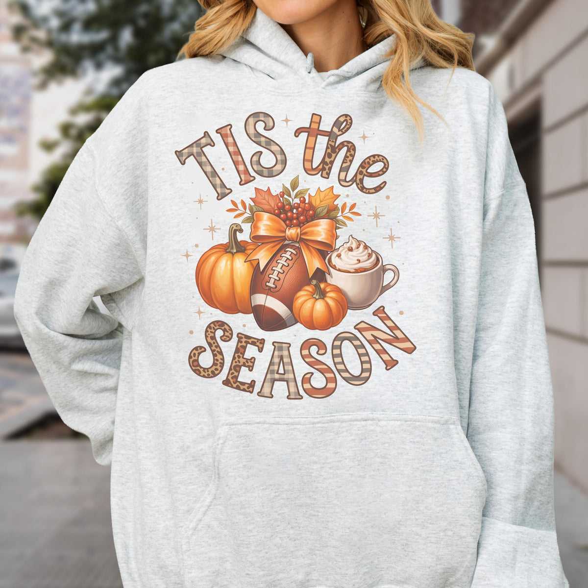 May include: Light gray hoodie with a fall-themed graphic. The design includes pumpkins, a football, a bow, and a coffee cup. The text reads "Tis the Season" in a mix of plaid and leopard print.