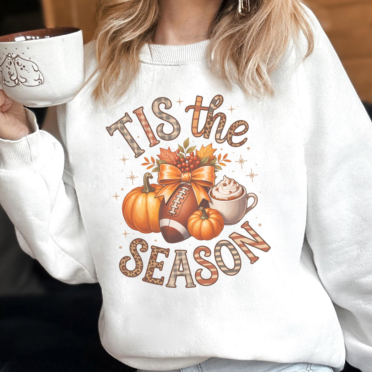 May include: White sweatshirt with a fall-themed graphic. The design includes pumpkins, a football, a bow, and a cup of coffee. The text "Tis the Season" is displayed in a decorative font, perfect for autumn.