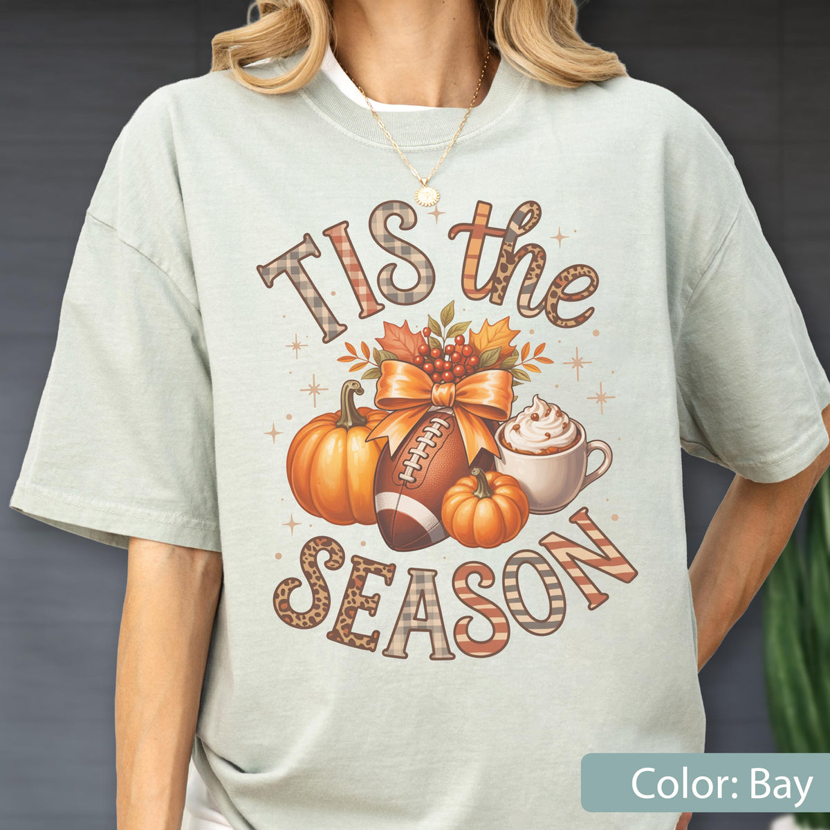 May include: A light green t-shirt with a fall-themed graphic. The design includes pumpkins, a football, a bow, and the text "TIS the SEASON". The shirt is a relaxed fit and has short sleeves.