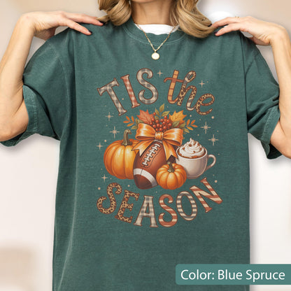 May include: Blue spruce t-shirt with a fall-themed graphic. The design includes pumpkins, a football, and a cup of whipped cream. The text reads "Tis the Season" and "Season" in a decorative font. The shirt is a relaxed fit.