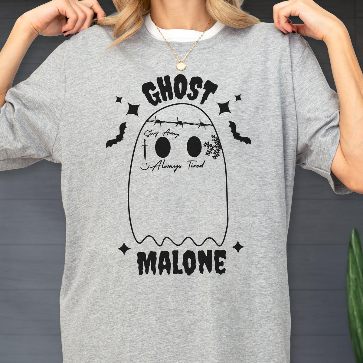 May include: A heather gray t-shirt featuring a black graphic of a ghost with barbed wire across its head, the text "GHOST MALONE," "Stay Away," and "Always Tired," along with a sword, a smiley face, a snowflake, bats, and four-pointed stars.