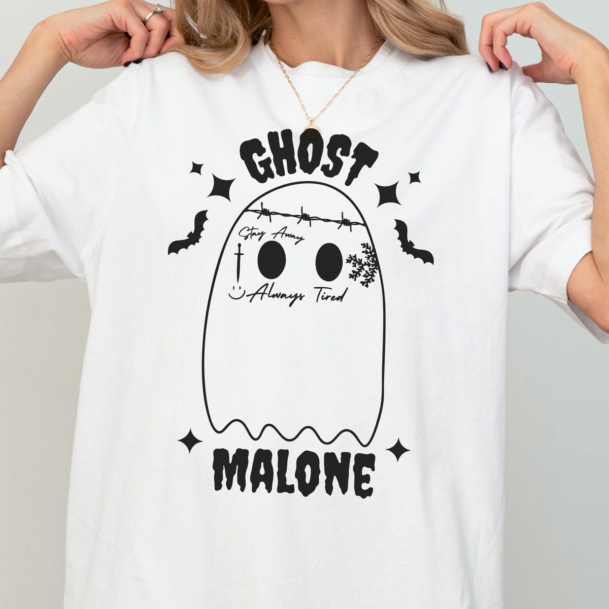 May include: A white short-sleeve t-shirt featuring a black graphic of a ghost with tattoos, including the text "GHOST MALONE", "Stay Away", and "Always Tired". The ghost design includes a barbed wire crown, a sword, a smiley face, and stars.
