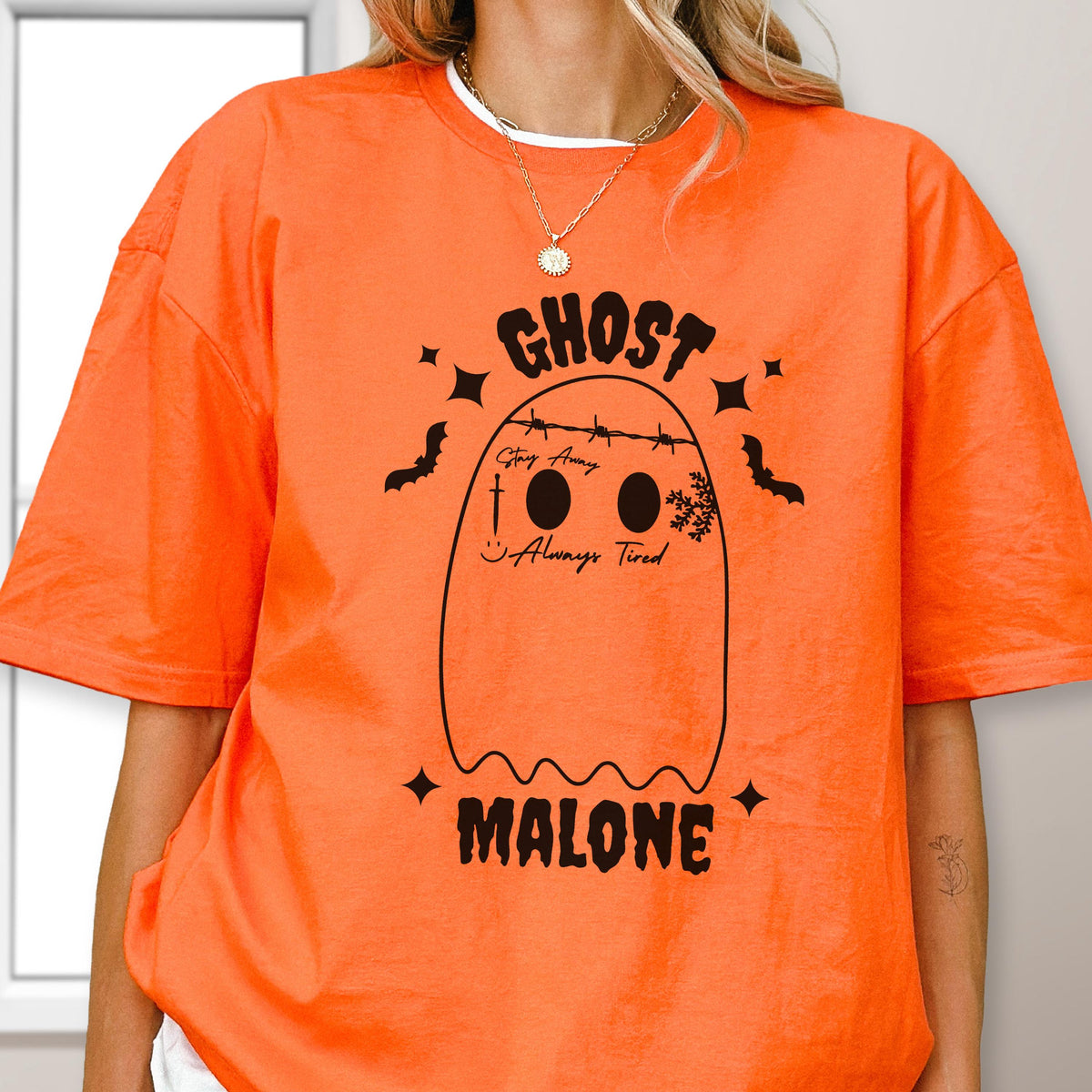 May include: An orange t-shirt featuring a black graphic of a ghost with barbed wire across its head, a sword, a snowflake, and the text "GHOST MALONE", "Stay Away", and "Always Tired" printed in black ink on the front of the garment.